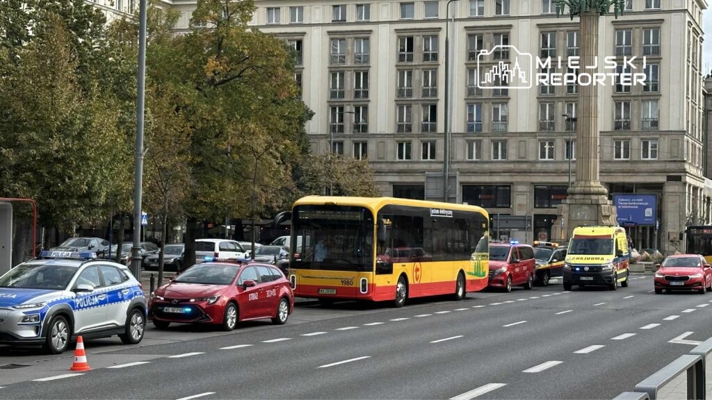 Ruch uliczny z żółtym autobusem, czerwonymi samochodami i karetką, w tle nowoczesny budynek i kolumna.
