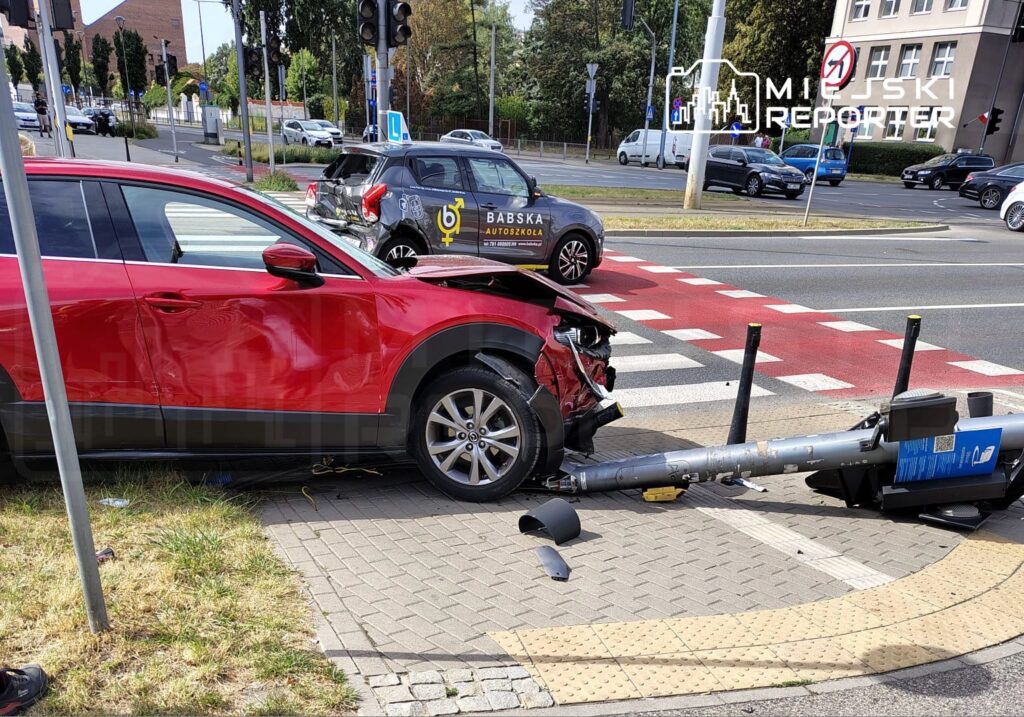 Czerwony SUV uderzył w latarnię na skrzyżowaniu, uszkadzając przód pojazdu, obok widoczny inny samochód.