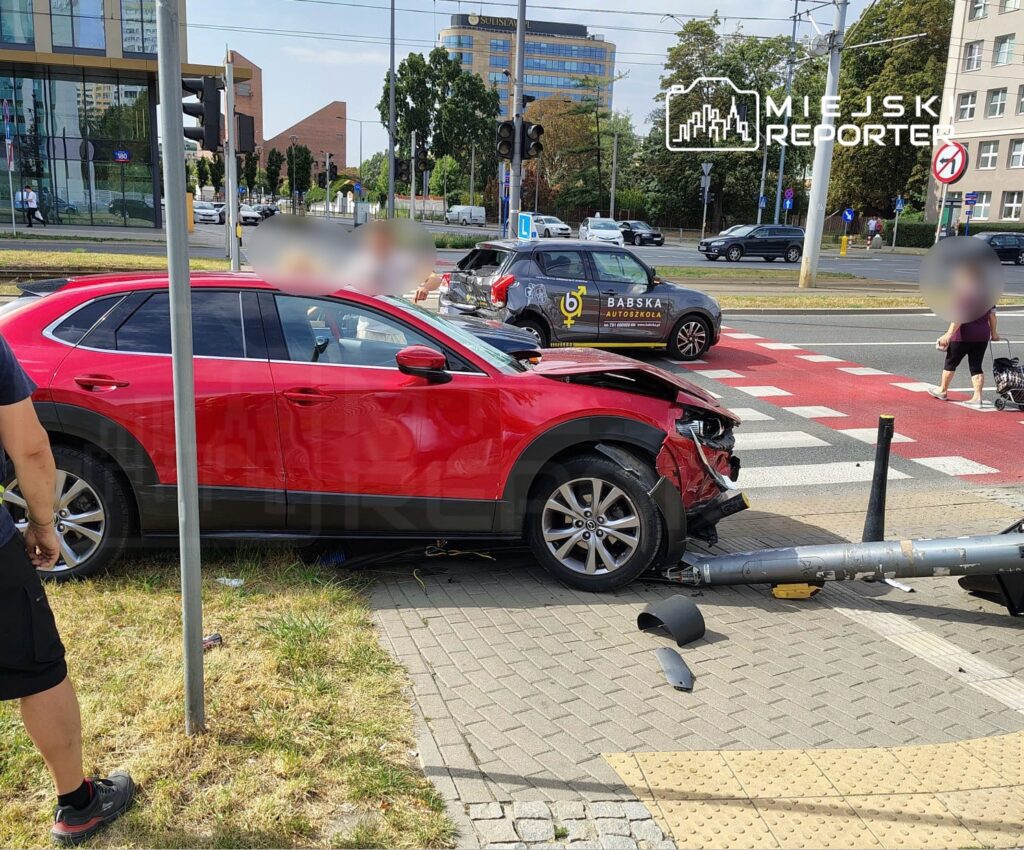 Czerwony samochód osobowy z uszkodzoną przednią częścią stoi przy przystanku, obok zniszczonego słupa.