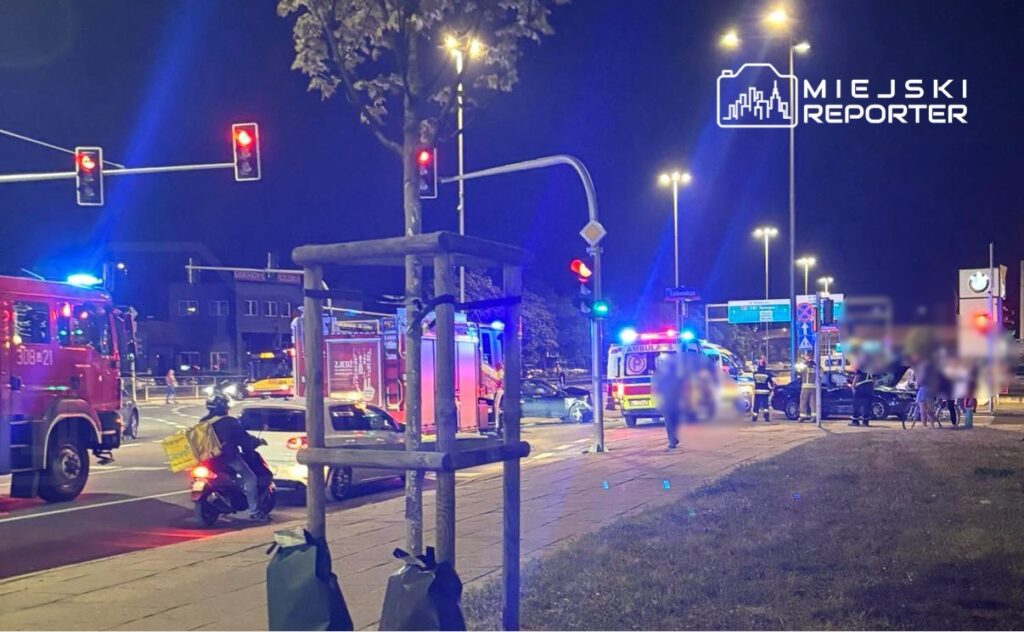 Na skrzyżowaniu z sygnalizacją świetlną stoją pojazdy służb ratunkowych, a w tle widać ludzi zbierających się wokół zdarzenia.