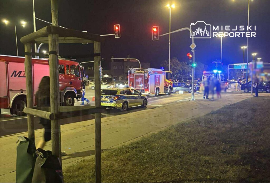 W nocy na skrzyżowaniu stoją pojazdy straży pożarnej i policji, a na drodze widać oznaczenia zabezpieczające.