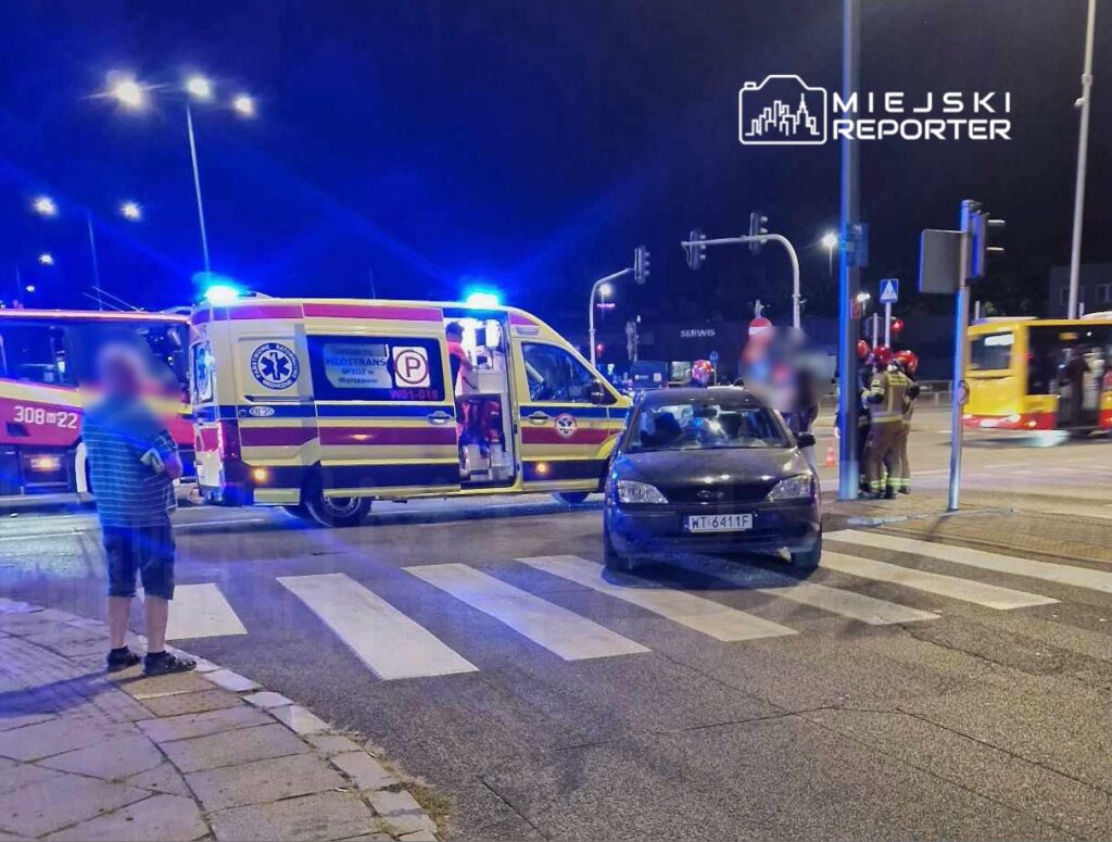 Na skrzyżowaniu, karetka z włączonymi sygnałami świetlnymi oraz strażacy interweniują przy czarnym samochodzie na przejściu dla pieszych.
