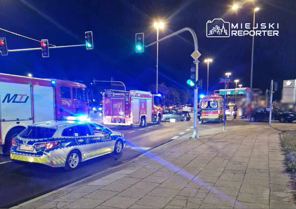 Na skrzyżowaniu w nocy zablokowane są pojazdy służb ratunkowych, w tym policja i straż pożarna, w związku z wypadkiem drogowym.