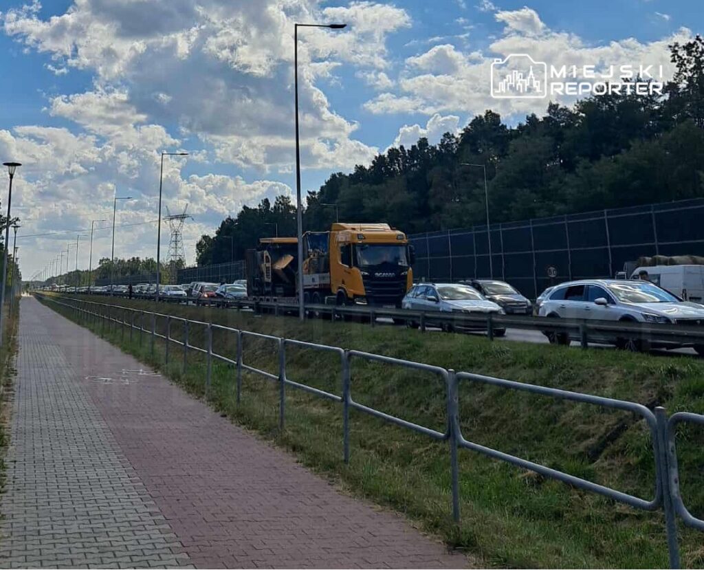 Ruchliwa droga z zatrzymanym ruchem, ciężarówka i samochody osobowe w kolejce, obok chodnik z kostki brukowej.