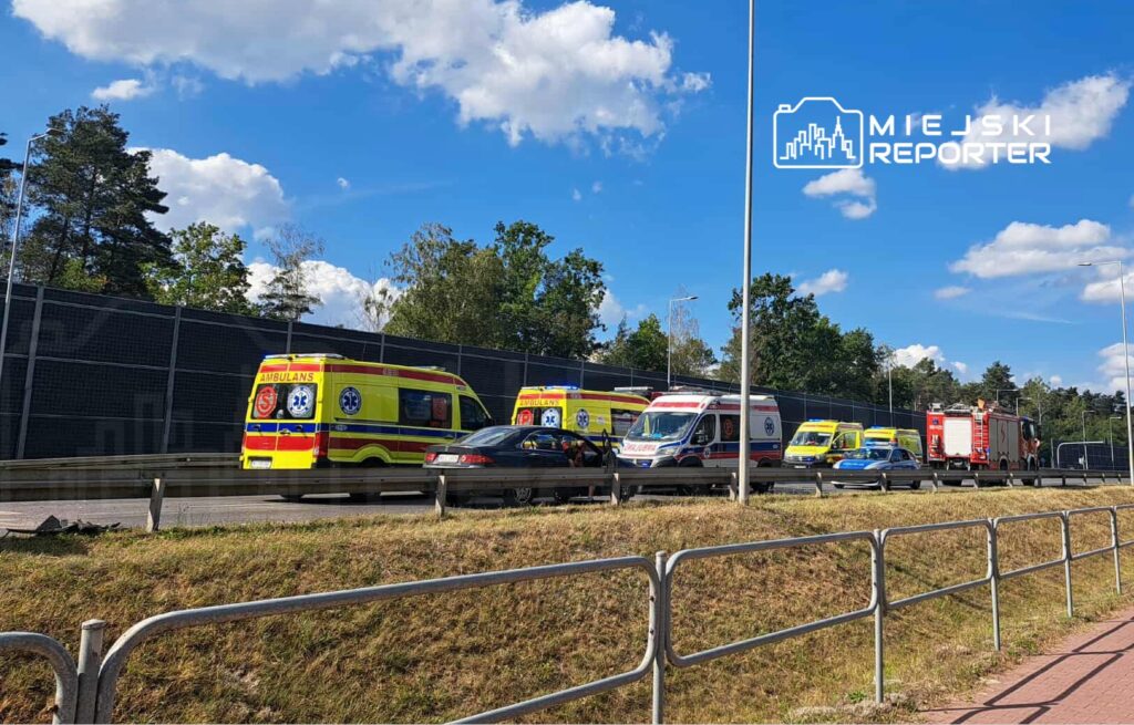 Karetki pogotowia oraz wóz strażacki stoją w zatorze drogowym na trasie, obok ogrodzenia i drzew.