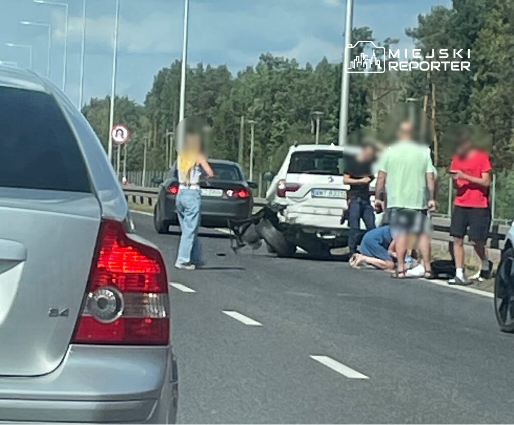 Na drodze stoi mężczyzna w krótkich spodniach, pomagający osobie leżącej na jezdni, obok grupka ludzi.