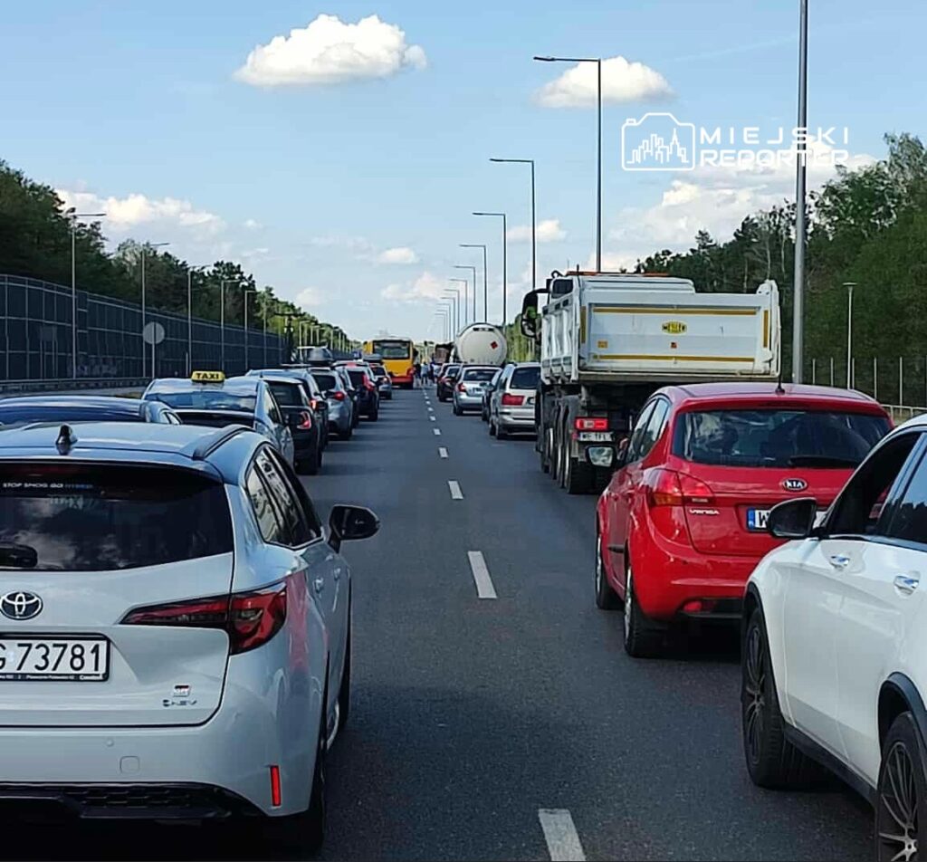 Ruch samochodowy w kierunku przeciwnym, z długim zatoriem na drodze, otoczony drzewami i ogrodzeniem.