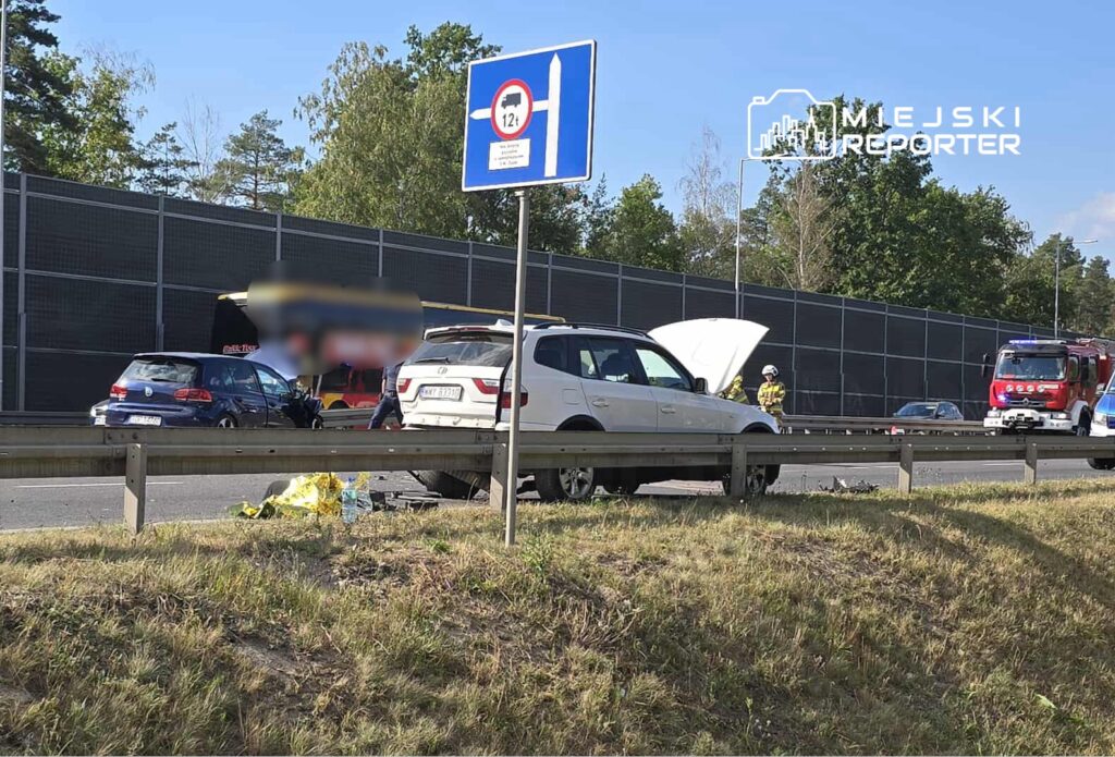 Biały SUV z uniesioną maską stoi przy drodze, obok interweniują strażacy i inne pojazdy. W tle widoczna sieć na ogrodzeniu.