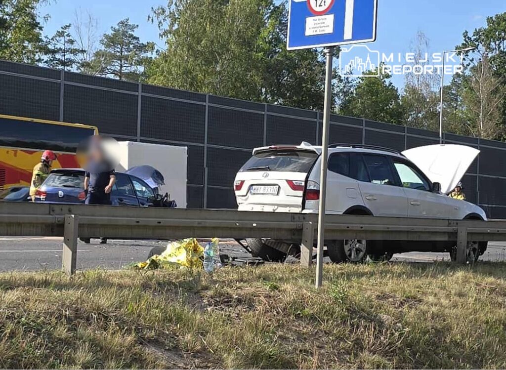 Na drodze, obok barierki, stoi biała terenówka z otwartą maską, obok niej pracują strażacy.
