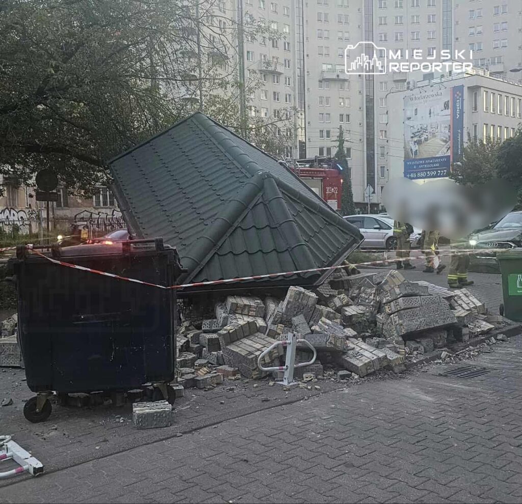 Zniszczona altana ogrodowa leży na boku obok kontenera na śmieci, otoczona gruzem i taśmą ostrzegawczą.