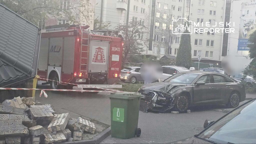W tle nowoczesne budynki, z przodu uszkodzony samochód osobowy oraz woz strażacki na ulicy, obok kontener na odpady.