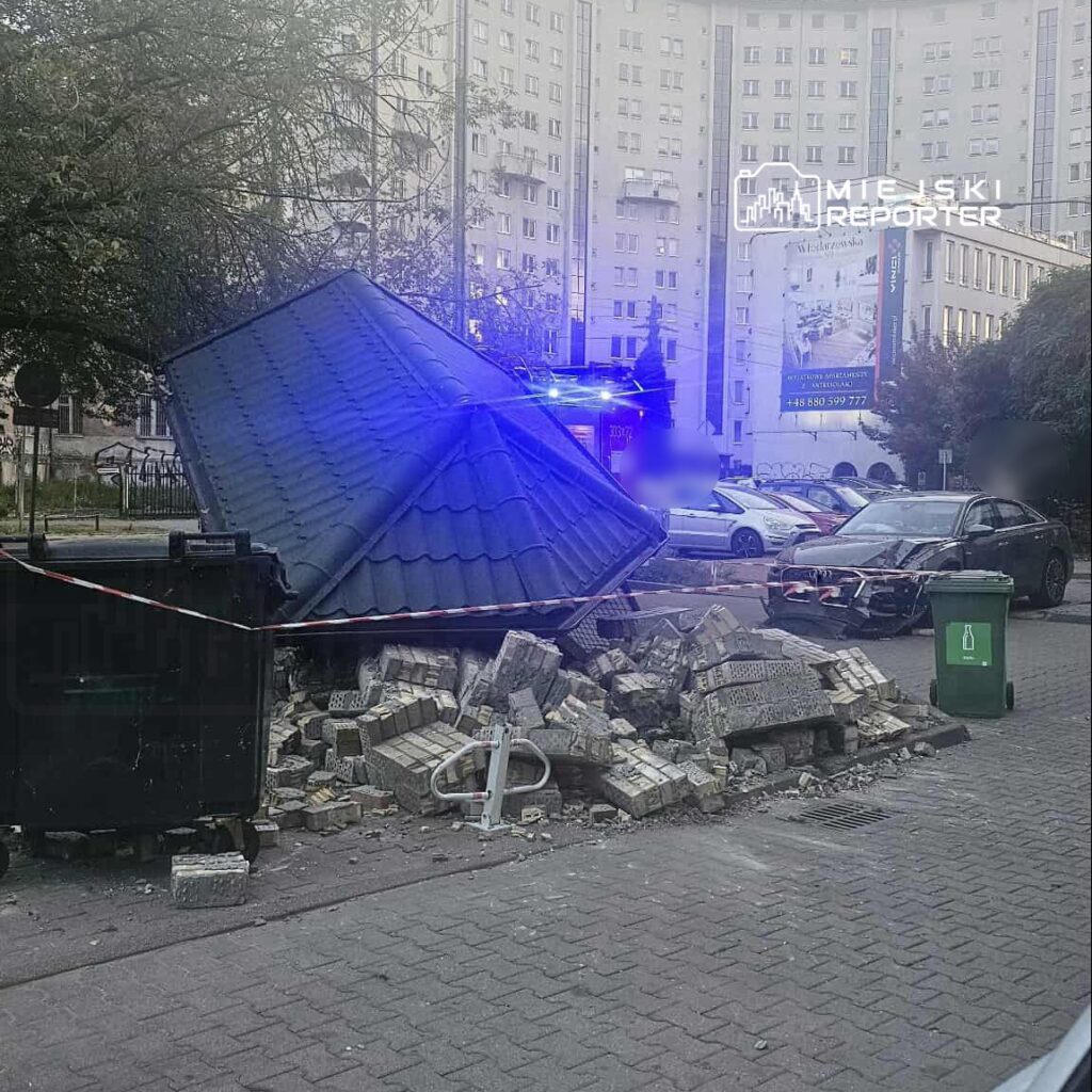 Zniszczony budynek z dachem leżącym na gruzach, obok stoją policyjne światła i uszkodzony samochód.
