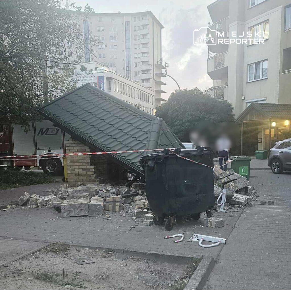 Zniszczona wiatka śmietnikowa przewrócona na bok, wokół leżą cegły i gruz, obok kontener na odpady.