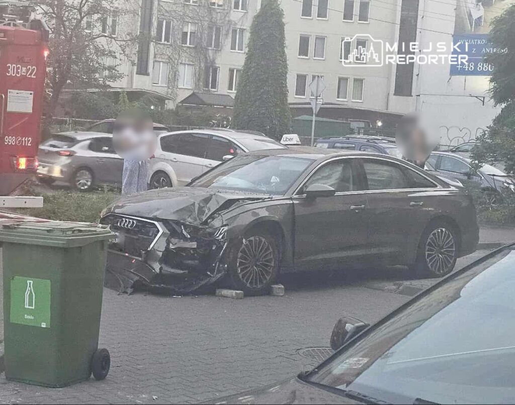 Uszkodzony czarny samochód osobowy zaparkowany na chodniku, obok straż pożarna i kilka osób obserwujących sytuację.