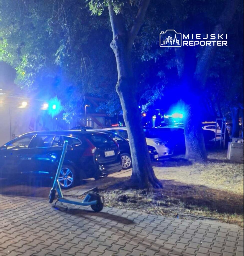 Nocna scena z niebieskimi światłami policyjnymi, zaparkowane samochody i elektryczna hulajnoga na chodniku.