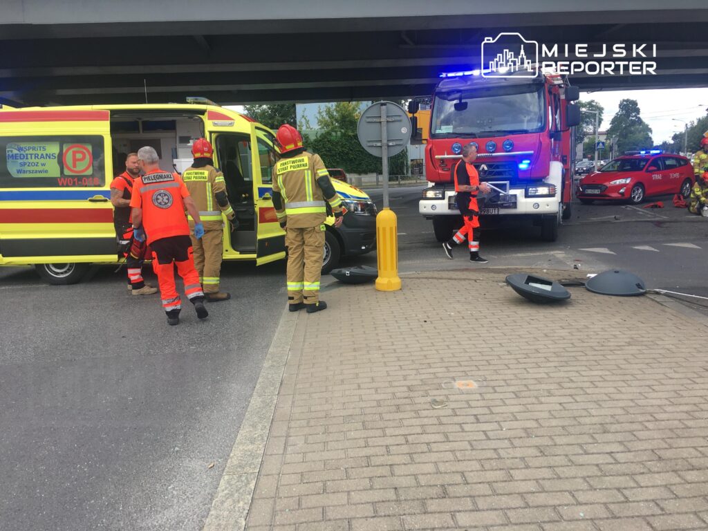 Ratownicy medyczni i strażacy pracują przy wypadku drogowym, obok ambulansu i wozu strażackiego na miejskiej ulicy.