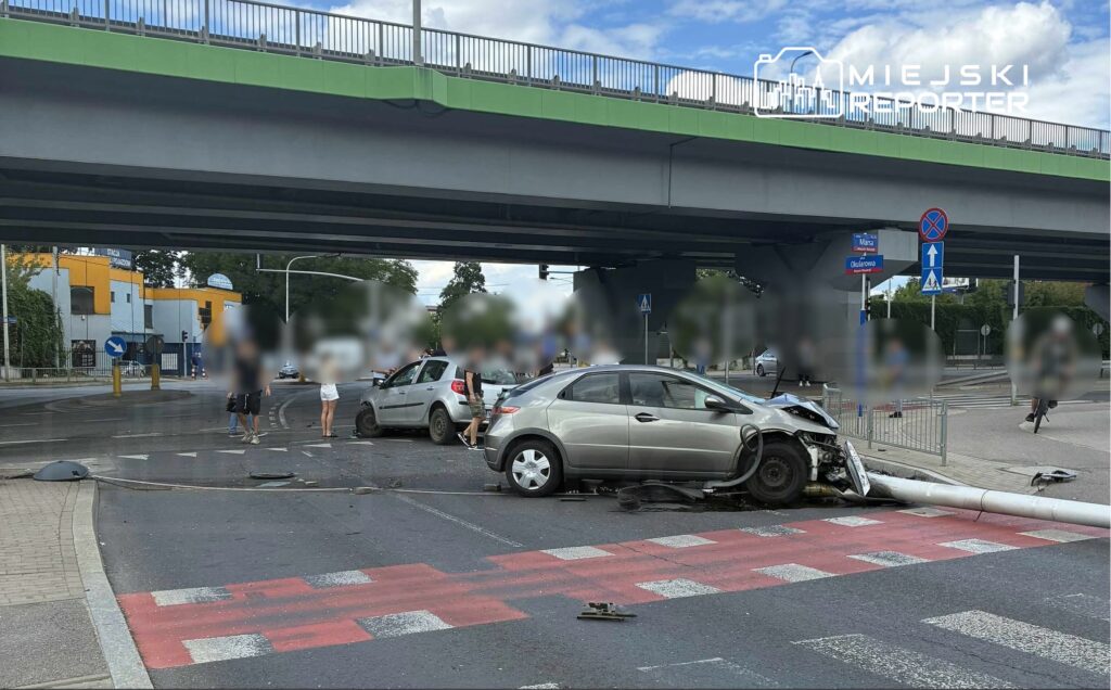 Dwa uszkodzone samochody na skrzyżowaniu, z fragmentami rozbitej infrastruktury leżącymi na drodze.