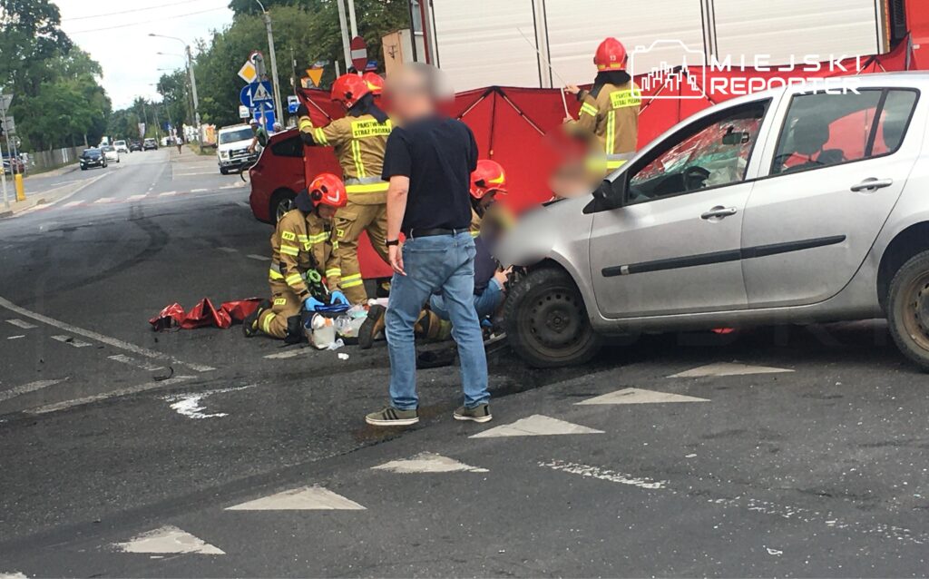 Grupa strażaków w hełmach udziela pomocy poszkodowanemu na jezdni obok uszkodzonego samochodu.