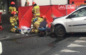 Ratownicy medyczni udzielają pomocy poszkodowanemu na jezdni, obok uszkodzonego samochodu, za czerwonym parawanem.