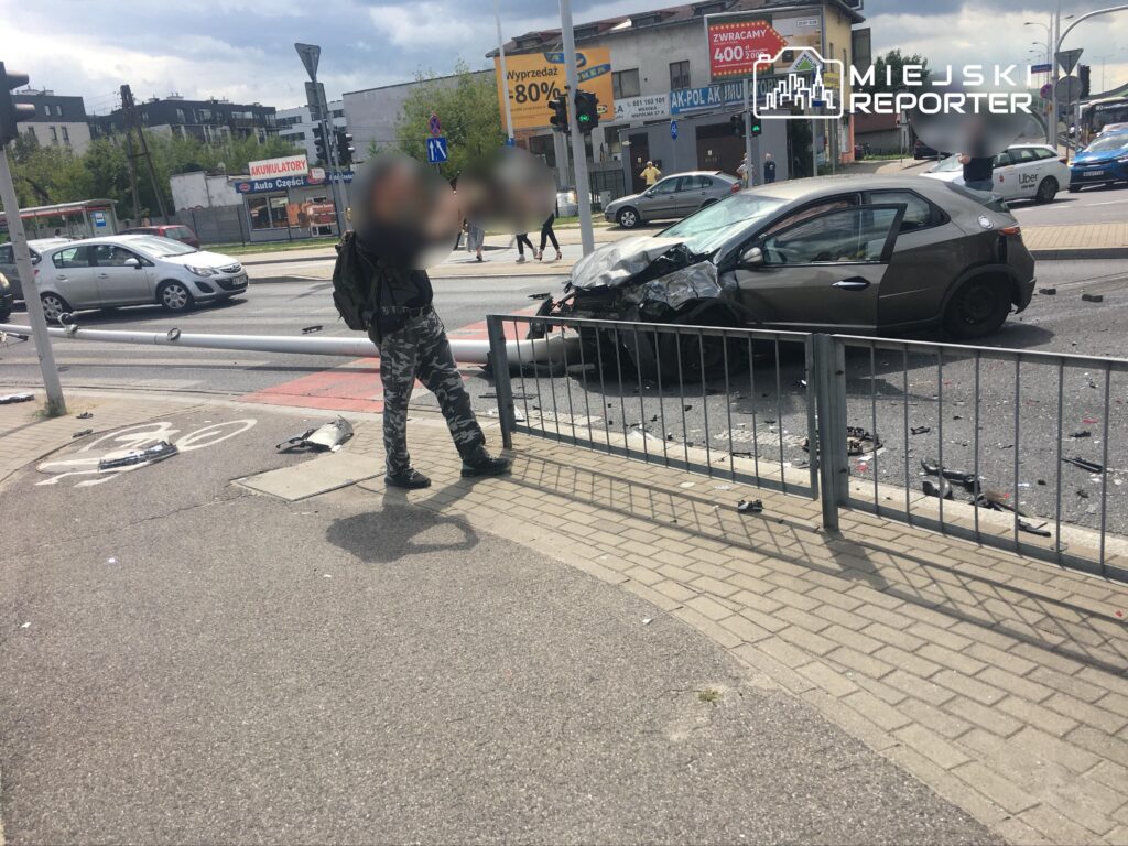 Mężczyzna w kamuflażowych spodniach obserwuje wypadek samochodowy na skrzyżowaniu, wokół leżą fragmenty pojazdów.