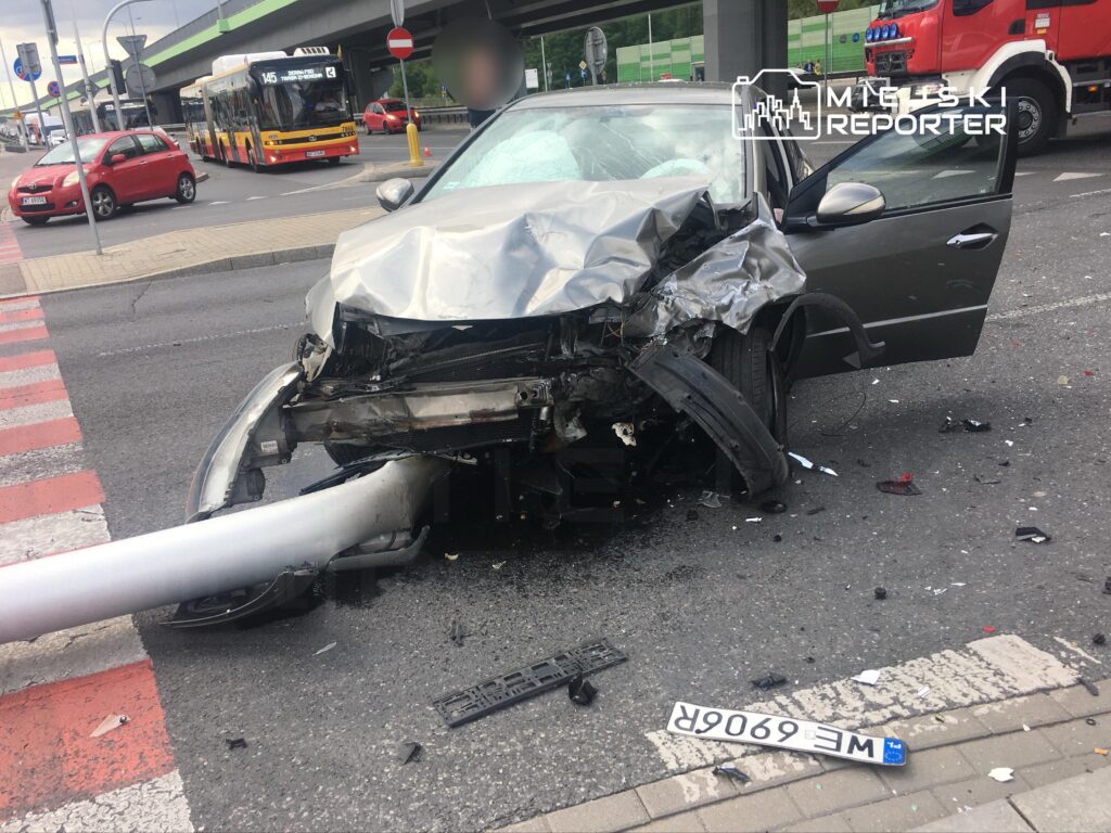 Zniszczony samochód osobowy uderzony w latarnię, porozrzucane elementy na jezdni, w tle przejeżdżający autobus.