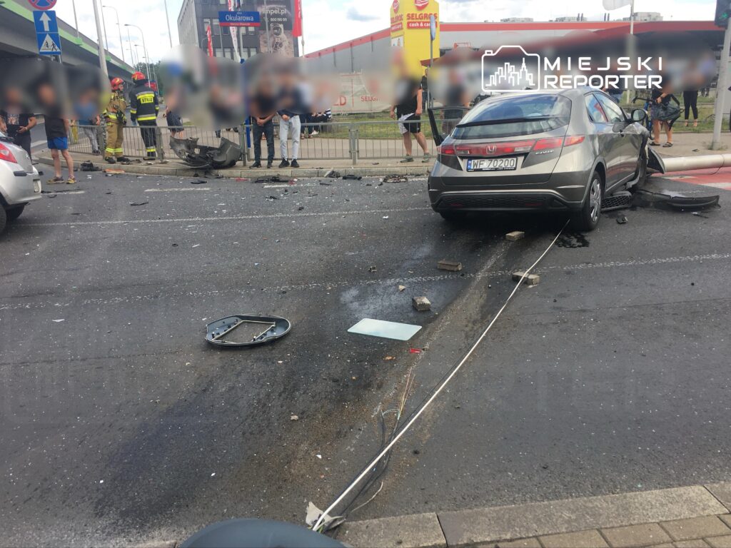 Zdemolowany samochód osobowy leży na drodze, otoczony przez gruz i fragmenty szkła, obok stoją świadkowie zdarzenia.