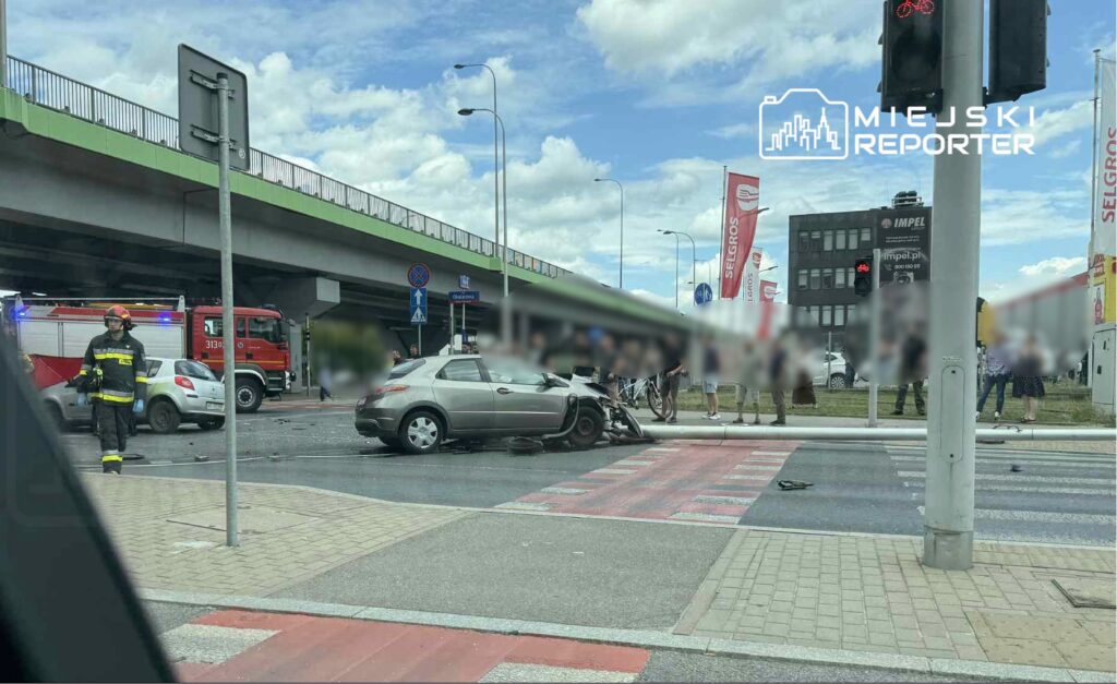 W miejscu wypadku samochodowego widać uszkodzony pojazd oraz strażaka z aparatem ochronnym, a w tle stoją inne samochody.