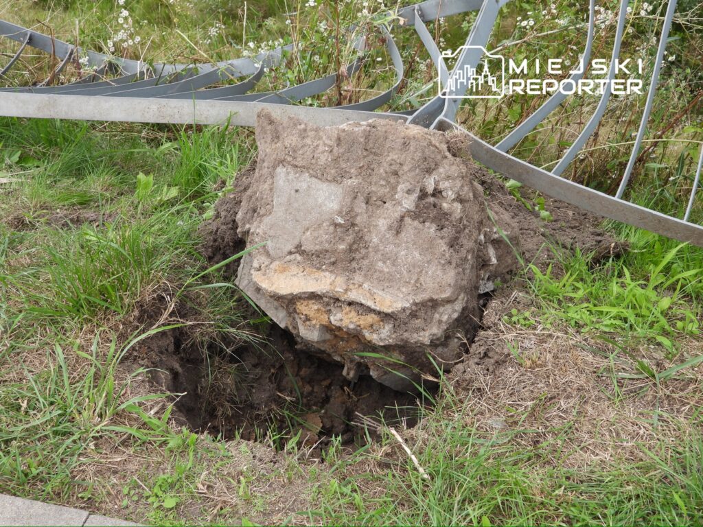Głaz wydobyty z ziemi, otoczony trawą i resztkami metalowej konstrukcji, w niewielkiej dziurze w ziemi.