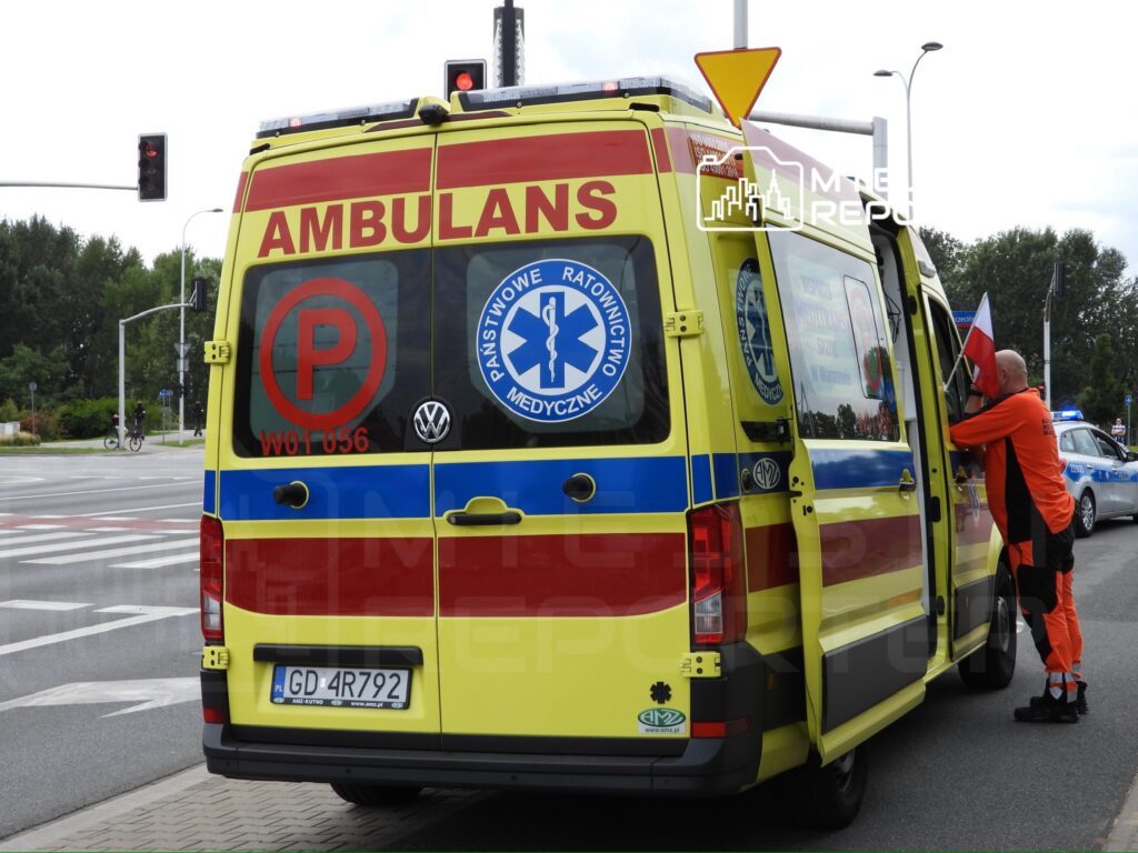 Ambulans z otwartymi drzwiami na ulicy, a ratownik w pomarańczowym stroju trzyma flagę.