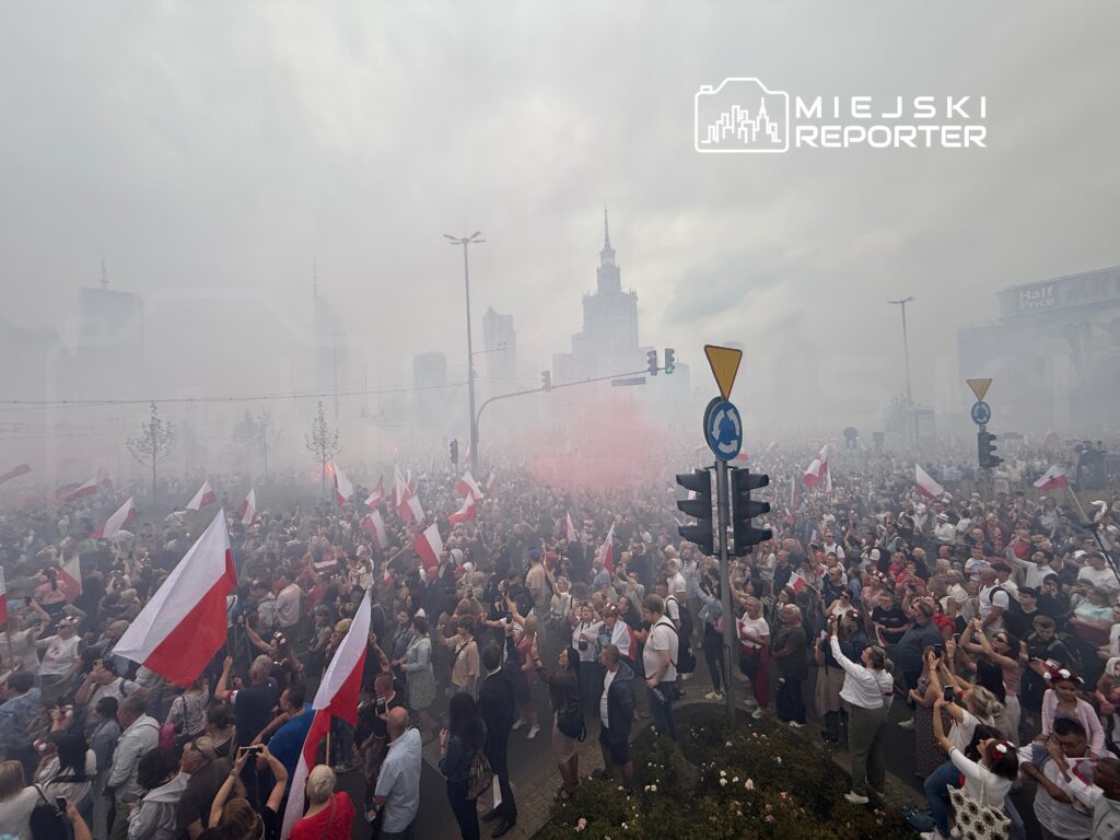 Tłum ludzi z flagami w barwach narodowych bawi się w gęstej mgle dymowej w miejskim otoczeniu.