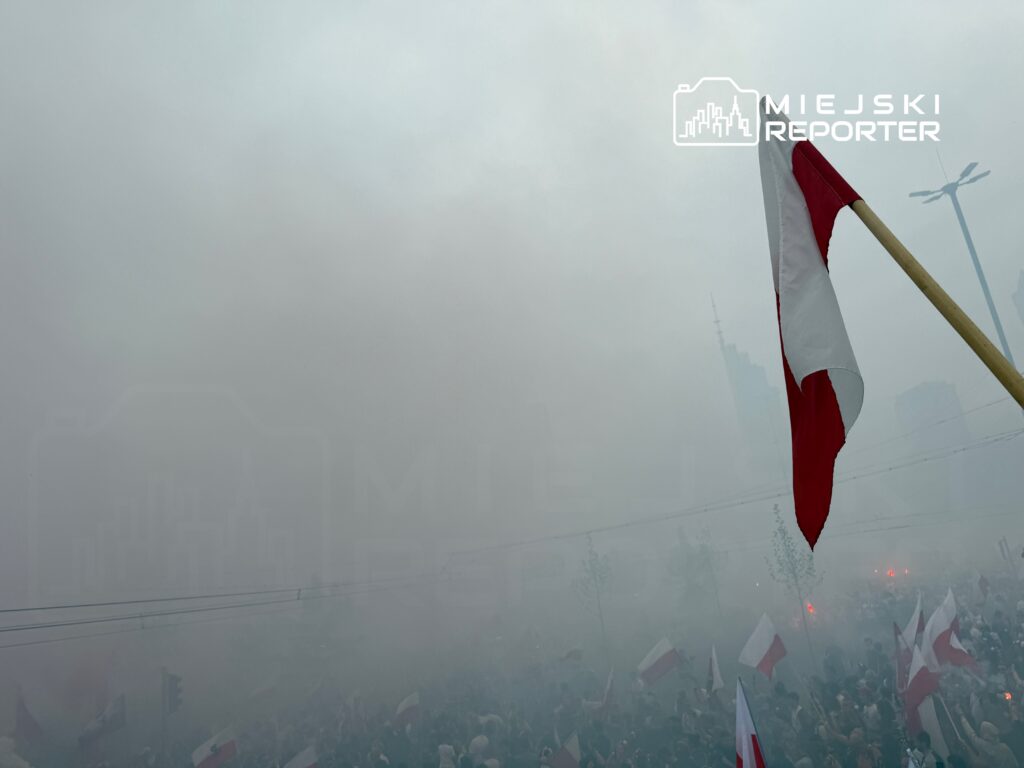 Tłum ludzi z flagami w chmurze dymu, w tle nowoczesne budynki.