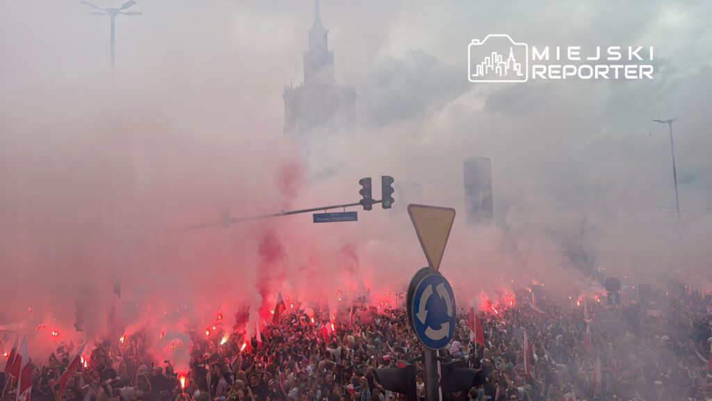 Tłum ludzi trzymających flagi, otoczony czerwonymi racami, w mglistym miejskim otoczeniu.