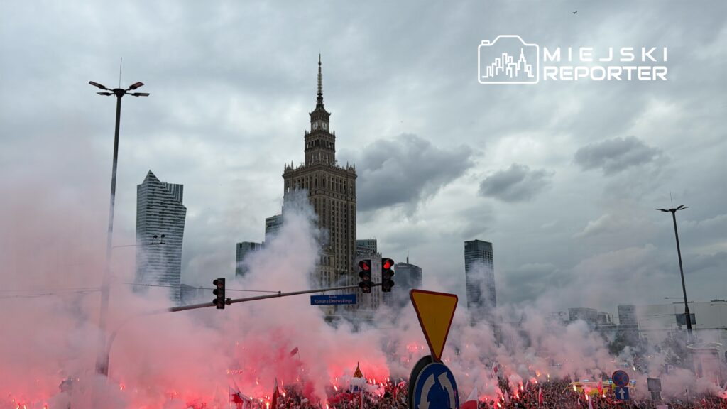 Tłum ludzi z flagami i racami świetlnymi zgromadzony na ulicy, w tle wieżowiec z zegarem.