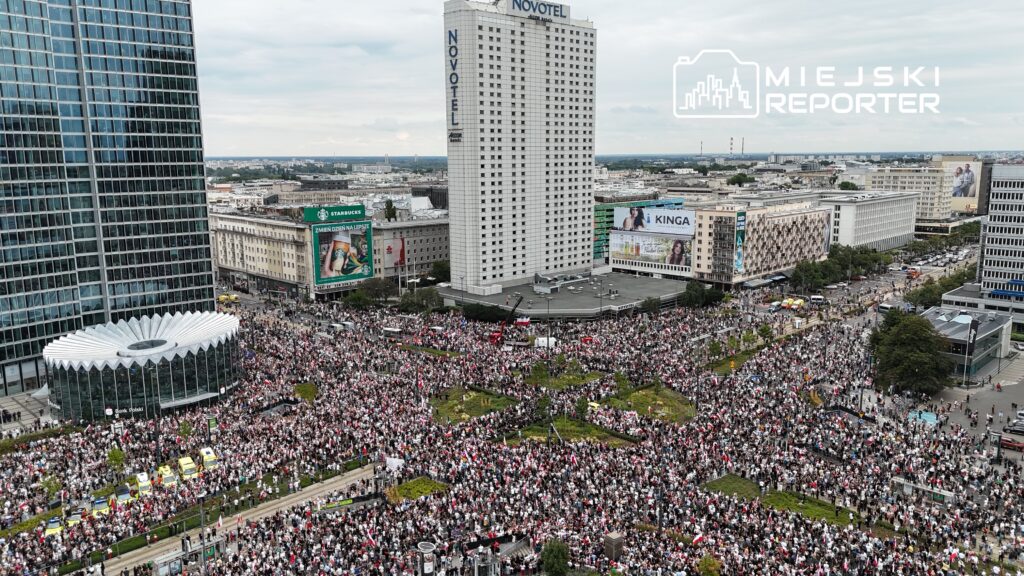 Tłum ludzi z flagami w dłoniach gromadzi się na placu przed nowoczesnym biurowcem w mieście.
