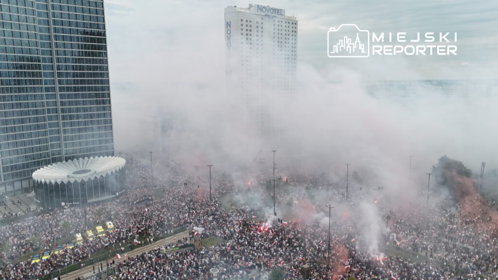 Tłum ludzi z flagami wznoszący okrzyki na ulicy, otoczony dymem z rac w pobliżu nowoczesnego budynku hotelowego.