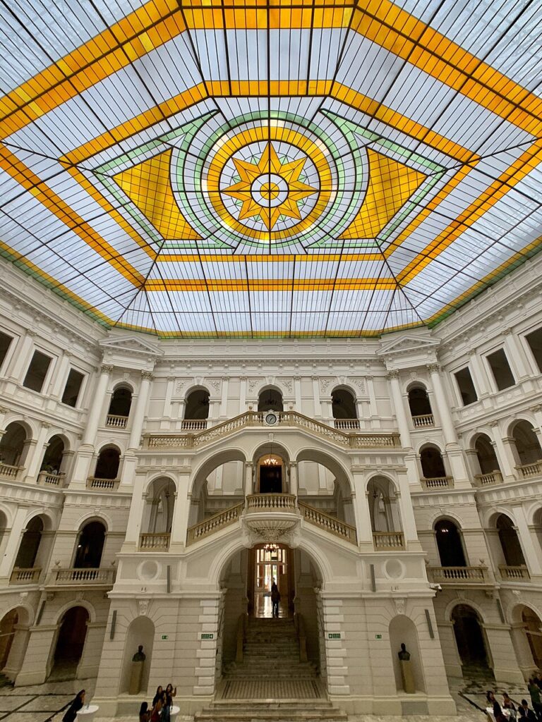 Wnętrze budynku z reprezentacyjną klatką schodową i witrażowym dachem w kolorach żółtym, zielonym i niebieskim.