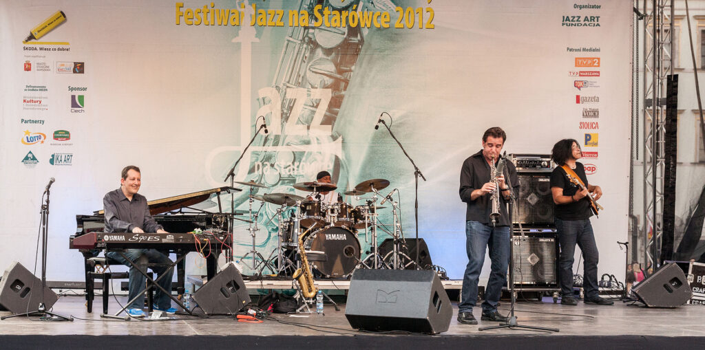 Muzyk grający na saksofonie podczas występu na festiwalu jazzowym, obok pianista i perkusista, w tle baner z nazwą wydarzenia.
