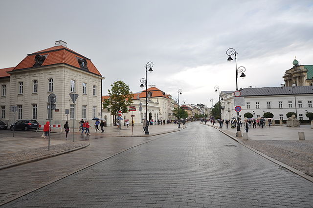 Grupa ludzi spaceruje po ulicy w miejskim otoczeniu z zabytkowymi budynkami i latarniami.