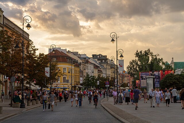 Tłum ludzi spacerujących po ulicy z kolorowymi kamienicami w tle, w blasku zachodzącego słońca.