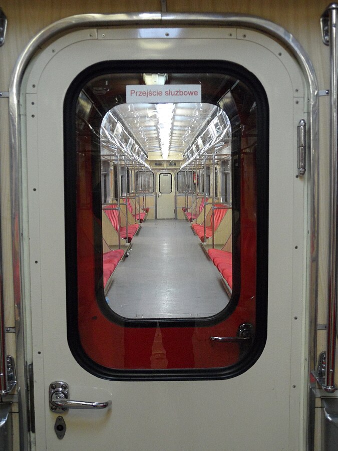 Pusty wagon metra z czerwonymi siedzeniami i widokiem na przeszklony korytarz. Na drzwiach widoczny napis: "Przejście służbowe".