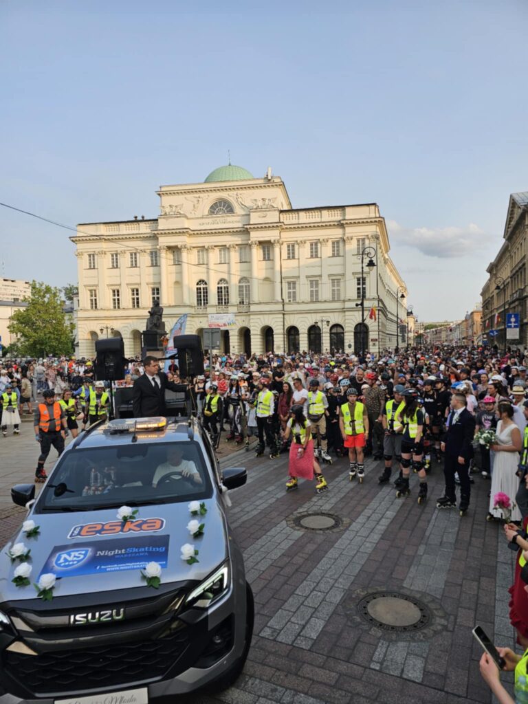 Mężczyzna w garniturze przemawia z samochodu wśród tłumu uczestników wydarzenia na ulicy.