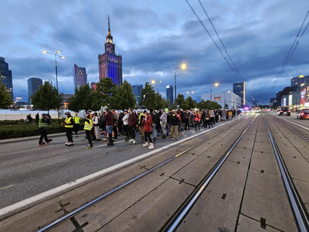 Grupa osób na rolkach czeka na sygnał do startu na ulicy, w tle widoczny nowoczesny wieżowiec.