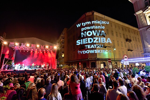 Tłum muzyków grających na scenie przed tłumem ludzi, na budynku widoczny napis: "W tym miejscu powstanie NOWA SIEDZIBA TEATRU ŻYDOWSKIEGO".