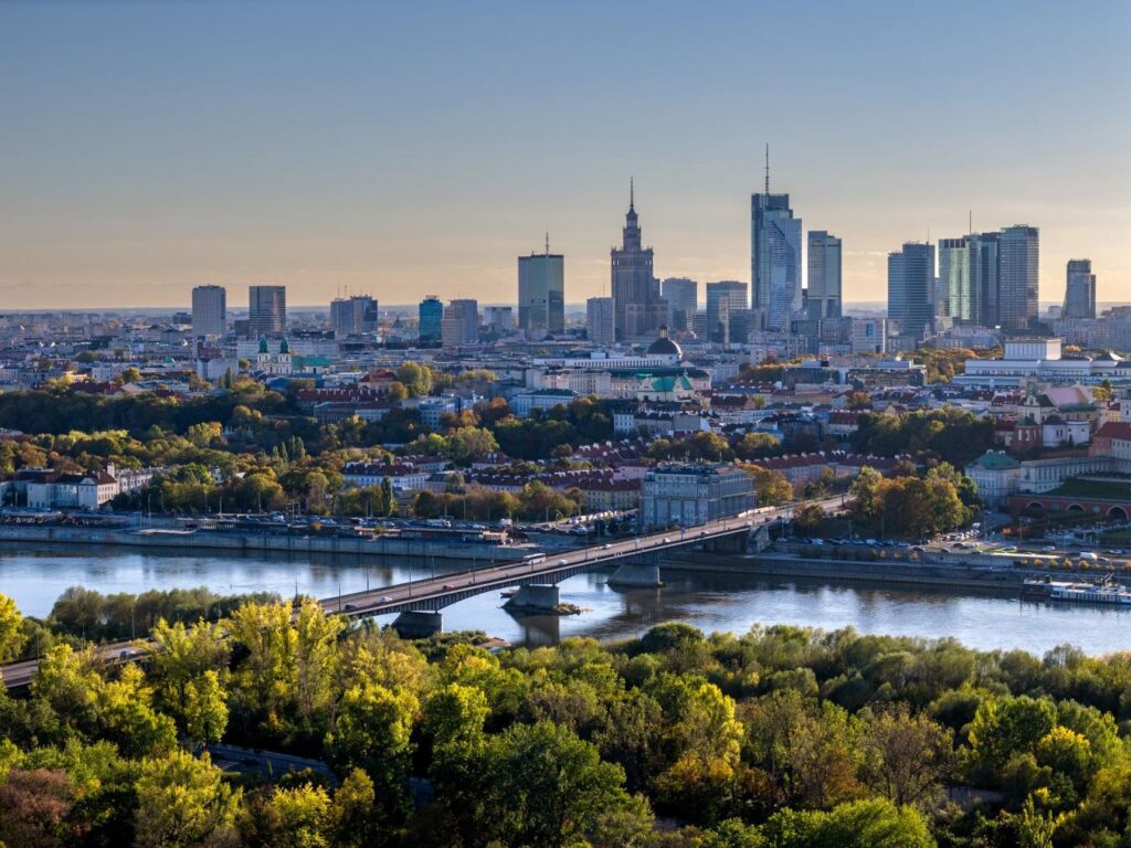 Warszawska panorama z nowoczesnymi wieżowcami w tle, rzeka Wisła i most łączący brzegi otoczone zielenią.