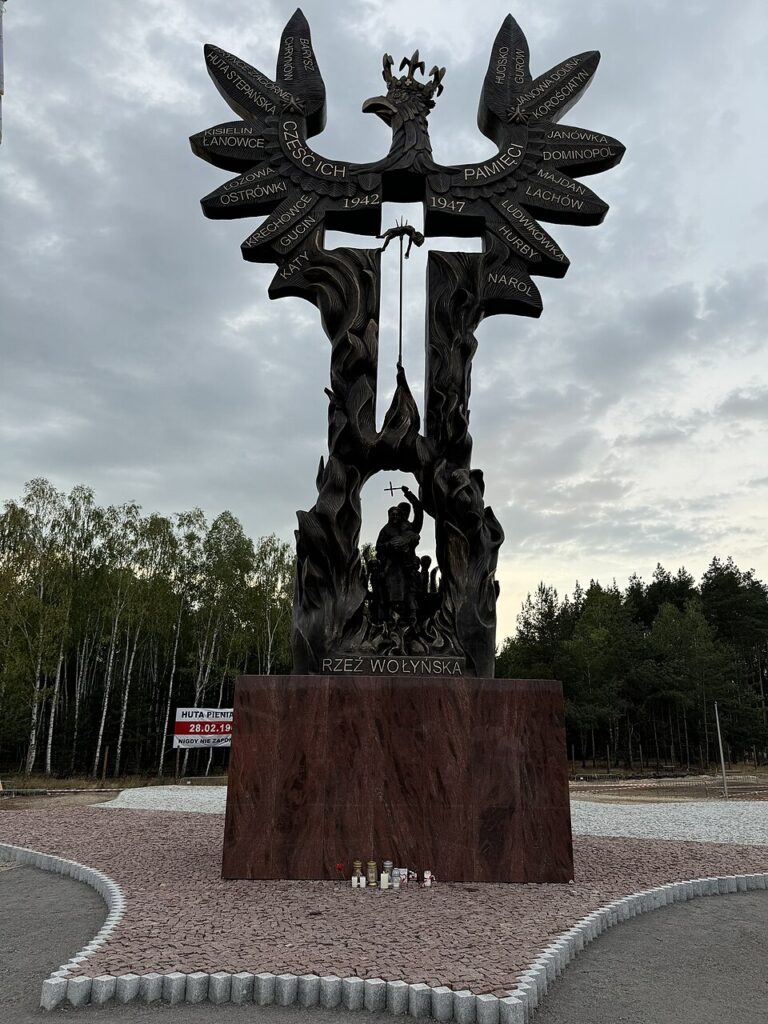 Pomnik upamiętniający ofiary Rzezi Wołyńskiej z widocznymi zniczami i kwiatami na tle lasu.