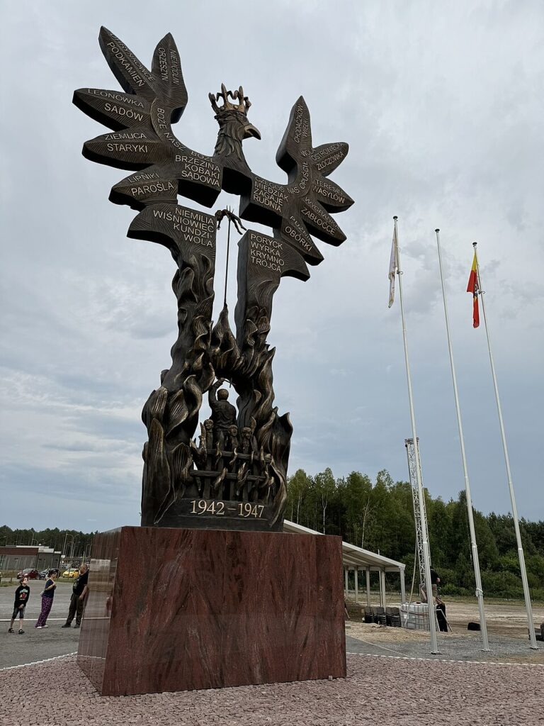 Pomnik w kształcie orła z nazwami miejscowości na skrzydłach, otoczony drzewami i flagami. W pobliżu stoją ludzie.