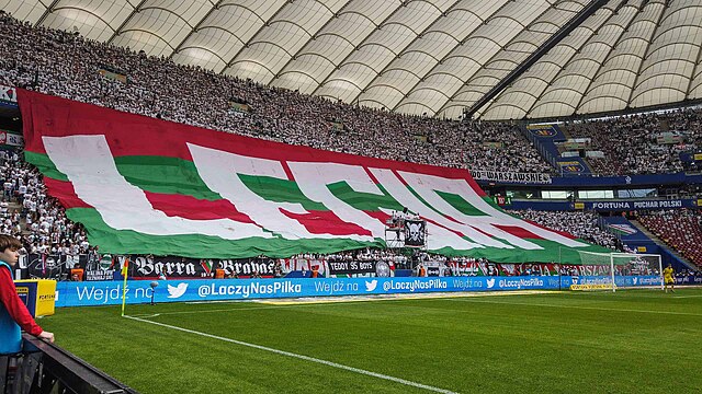 Kibice na stadionie prezentują dużą flagę z napisem "LECHIA" w barwach klubowych.