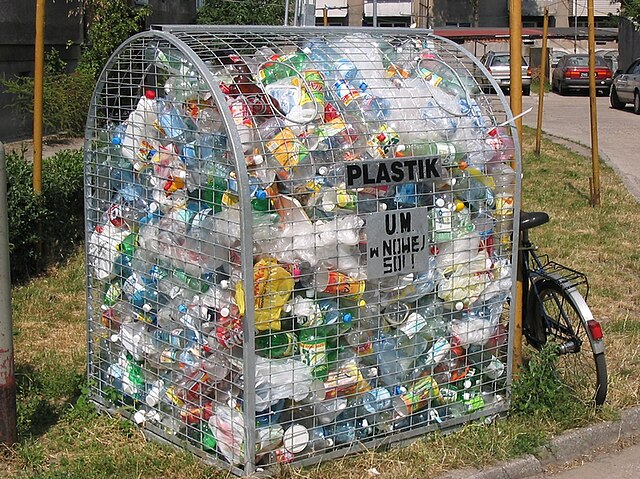 Kosz na plastikowe odpady wypełniony butelkami i opakowaniami, umieszczony na trawniku obok roweru.