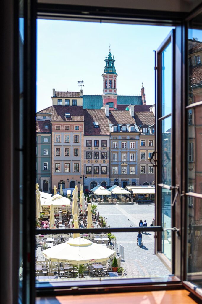 Widok z okna na rynek z kolorowymi kamienicami i parasolami przy stolikach w słoneczny dzień.