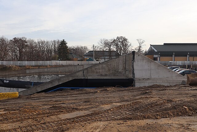 Betonowa konstrukcja z nachyloną rampą w trakcie budowy, otoczona ziemią i drzewami w tle.